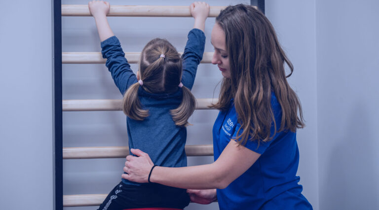 Zdjęcie przedstawia fizjoterapeutkę Karolinę Mierzejewską rehabilitującą małą dziewczynkę siedzącą na piłce przy drabinkach gimnastycznych.