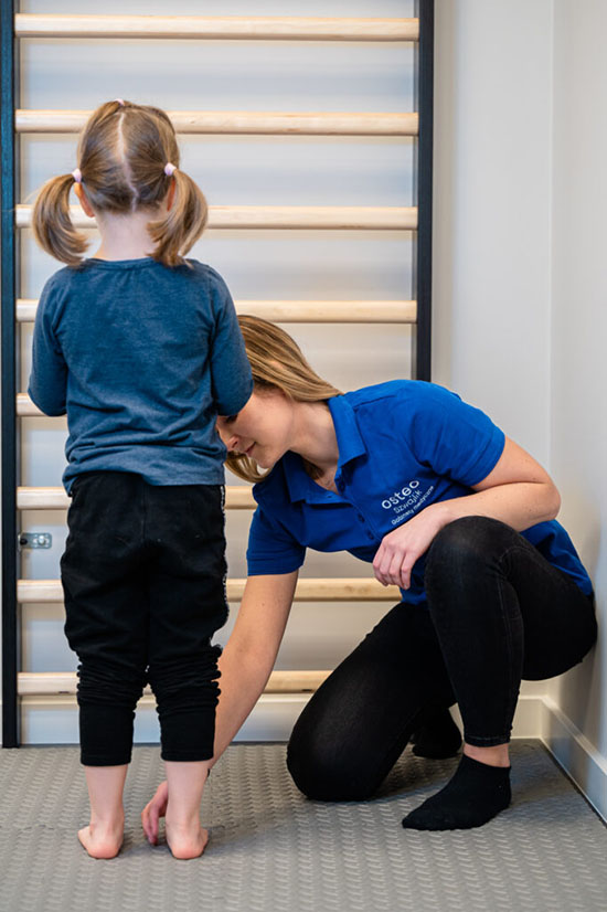 Zdjęcie przedstawia rehabilitacje wad stóp u dziecka Karolina Mierzejewska. Dziecko stoi na materacu przed drabinkami a fizjoterapeutka diagnozuje wadę postawy.